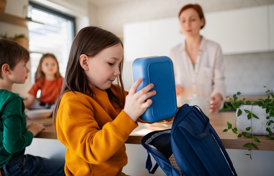girl packing lunch box into backpack while in kitchen at home with family