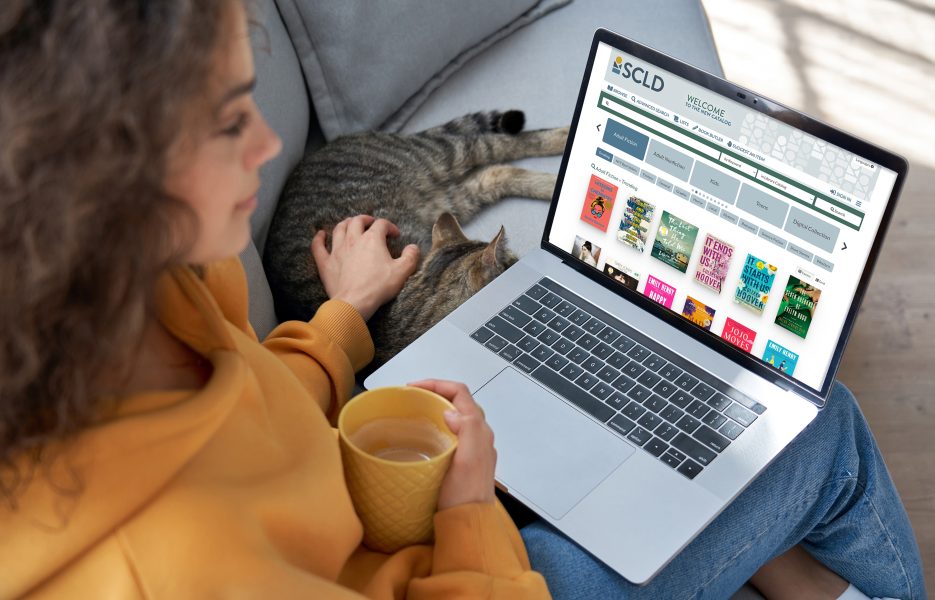 Woman looking at the library catalog on her laptop while sitting on a couch, holding a mug, and petting a cat