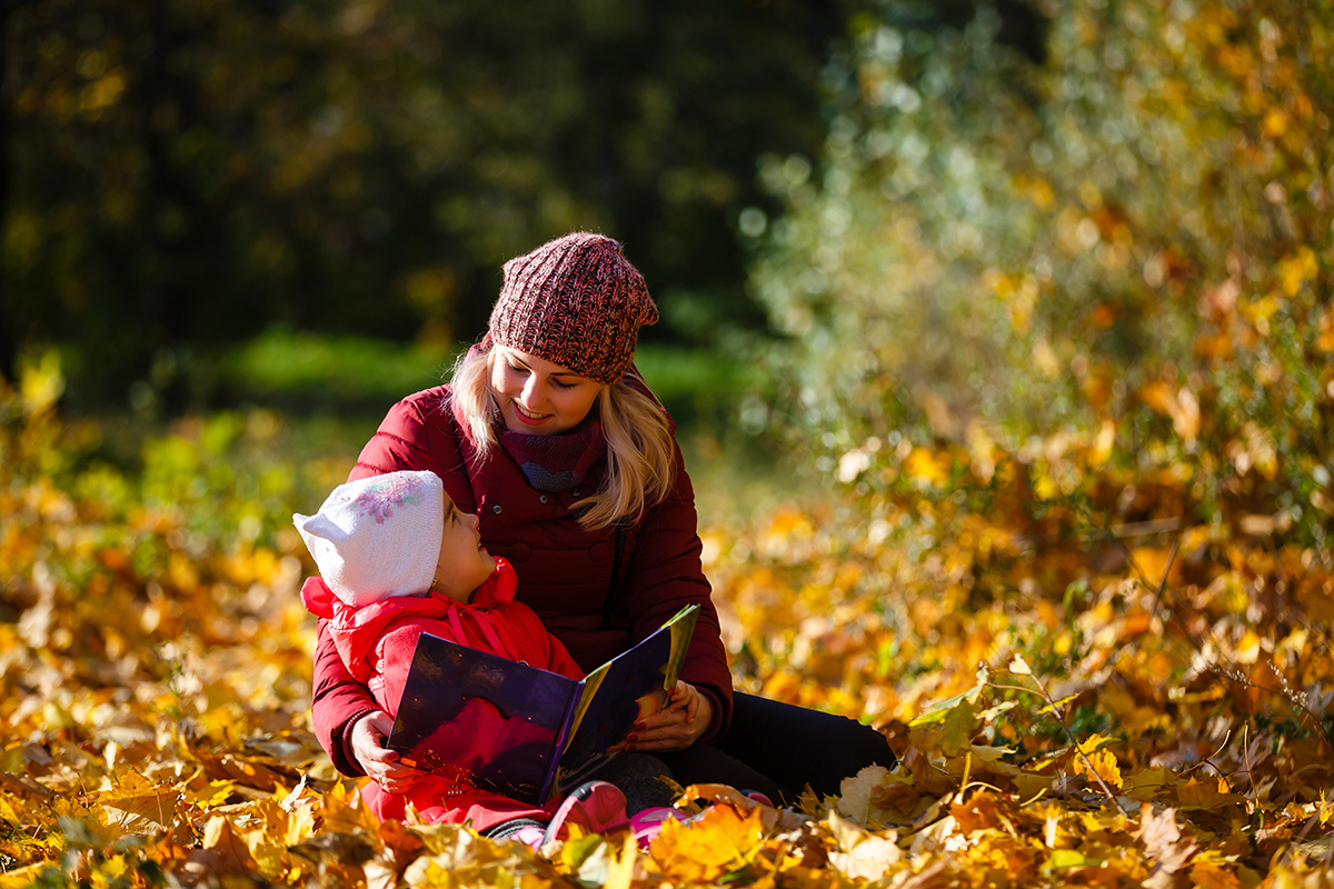 Mother and daughter enjoying reading a book together relaxing in the autumn leaves at the park