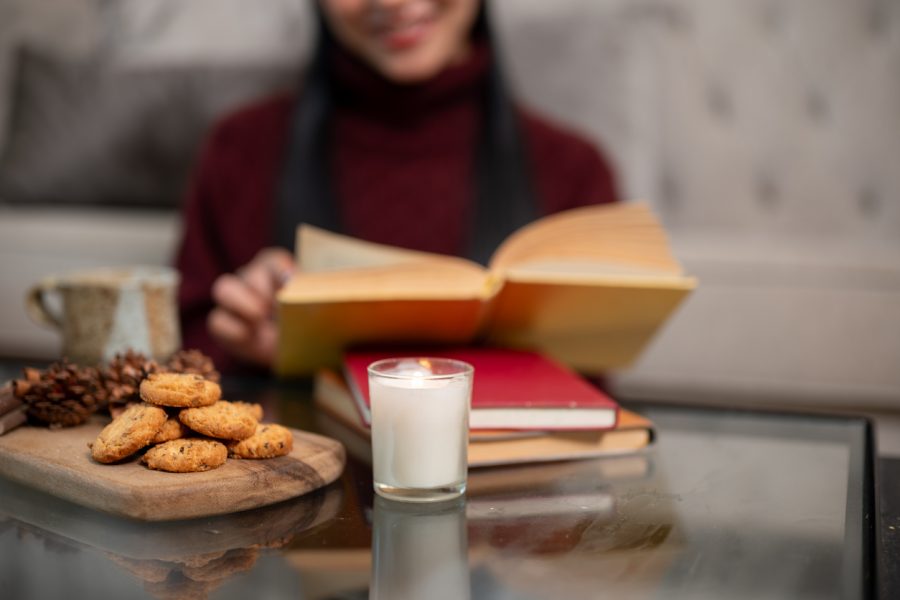 A wooden plate of cookies, a lit candle, stacked books, and a coffee mug on a coffee table. In the blurred background, a woman sits reading, creating a cozy Christmas at home atmosphere.