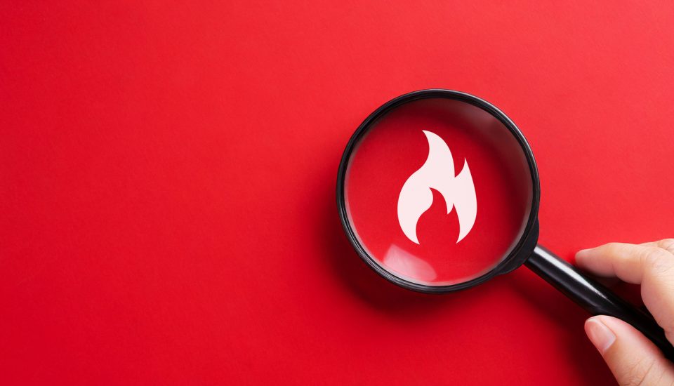 Hand holding a magnifying glass focused on a flame icon with a red background.